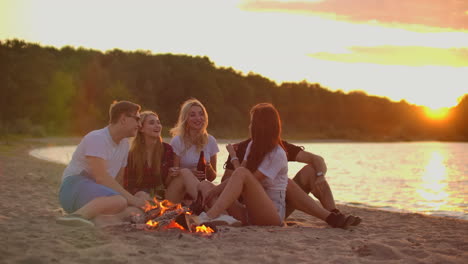 The-young-blonde-is-telling-a-funny-story-to-her-friends-around-bonfire-on-the-beach.-They-are-drinking-beer-at-sunset-and-enjoying-the-summer-evening-on-the-lake-coast.
